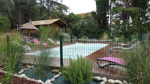 Un área de piscina con sillas de descanso, una estructura de techo de paja y un jardín cercado con rocas y plantas.