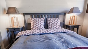 Una cama bien hecha con una colcha azul y roja con estampado, dos almohadas con diseño de copo de nieve y dos lámparas de noche sobre mesitas de madera.
