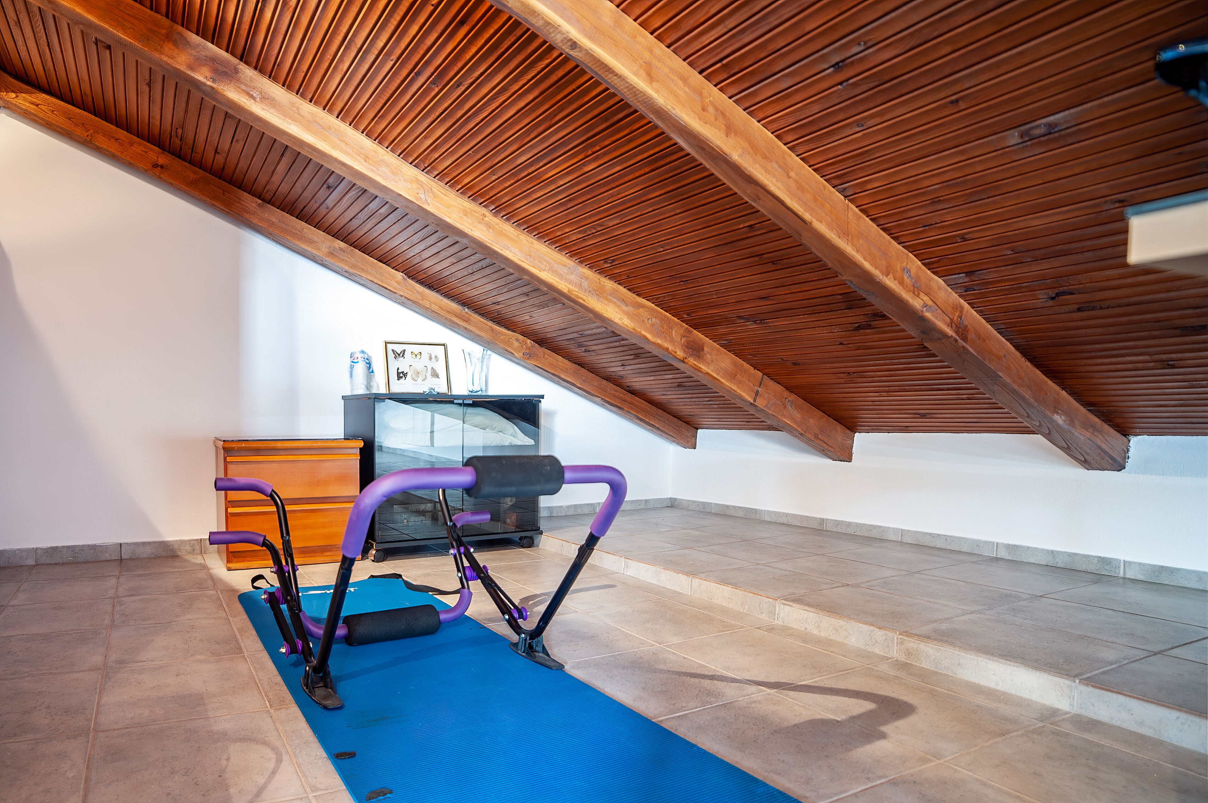 Una habitación en el ático con techo de madera, equipo de ejercicio, televisor y cómoda.