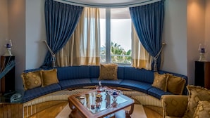 A curved blue sofa with patterned cushions, a glass coffee table with decorative items, and a view of greenery through large windows with blue and beige curtains. - Villa Serenity (Kymi-Aliveri)
