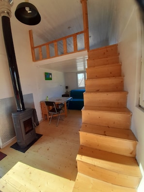 Una escalera de madera que lleva a un nivel superior, un horno de leña, una mesa de comedor con sillas y un sofá azul en una habitación con pisos de madera y paredes blancas.