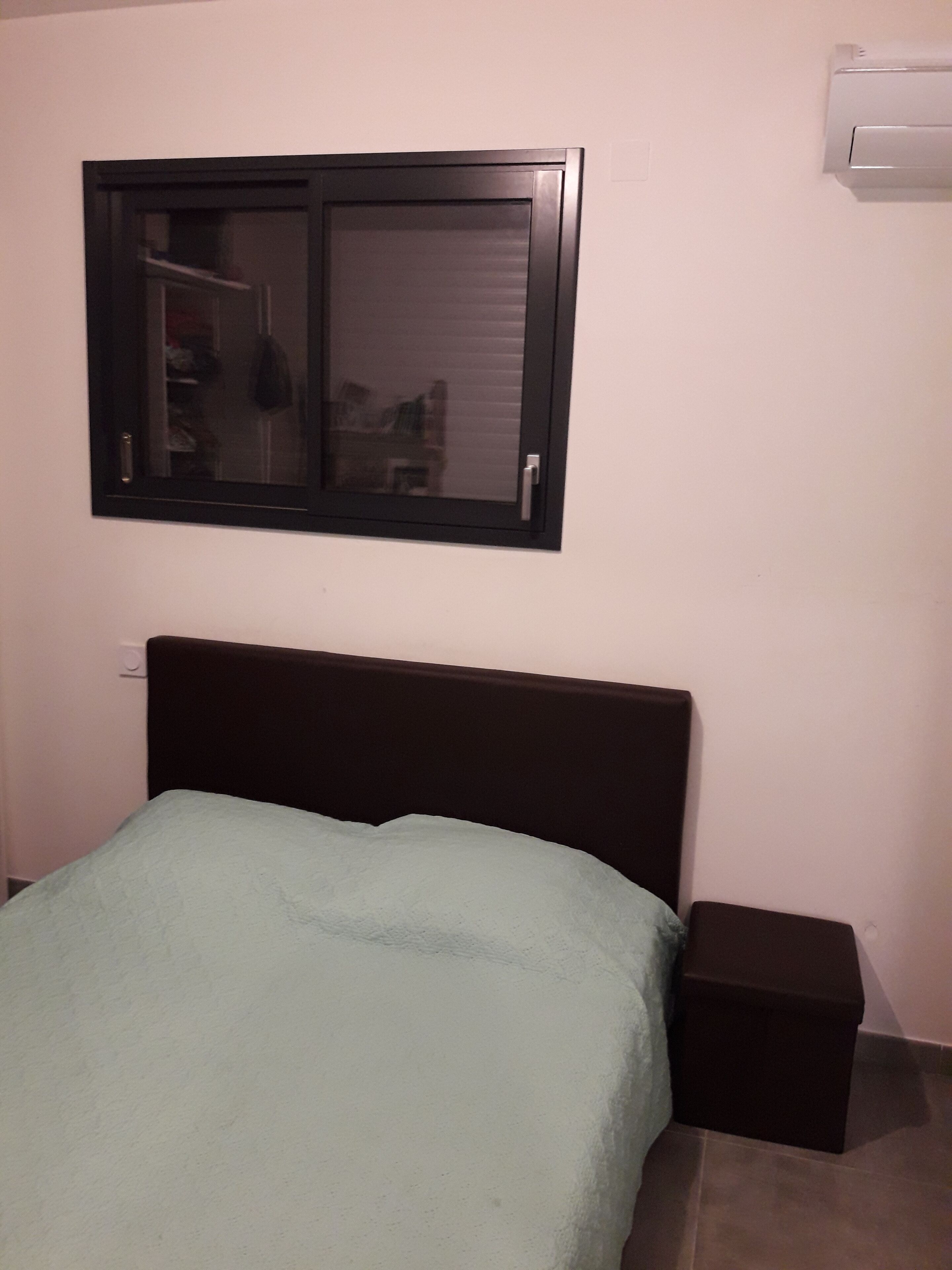 Een slaapkamer met een bed, nachtkastje, raam en een airconditioning.