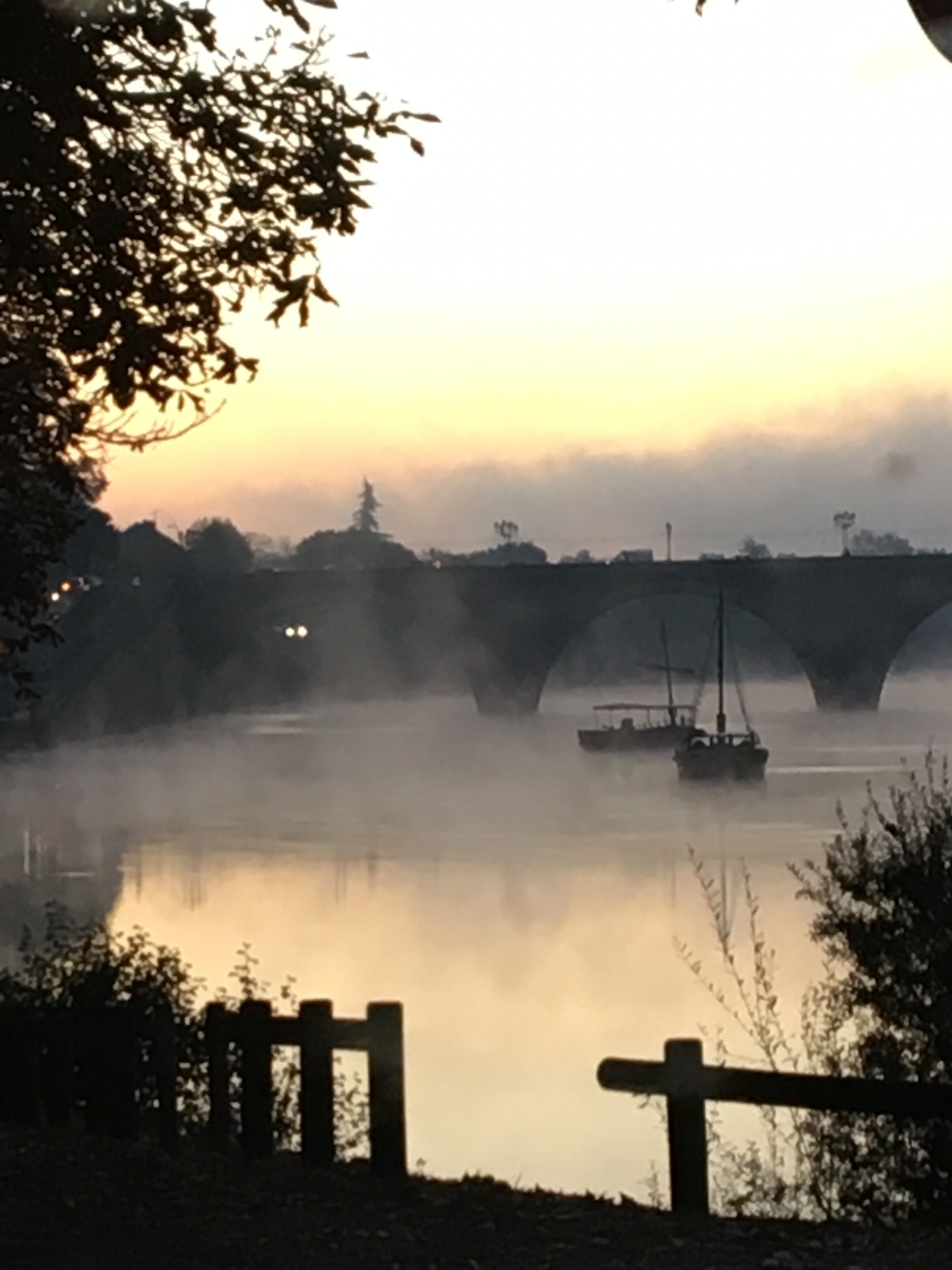 A bridge over a calm river with a boat in the distance, surrounded by mist and trees.