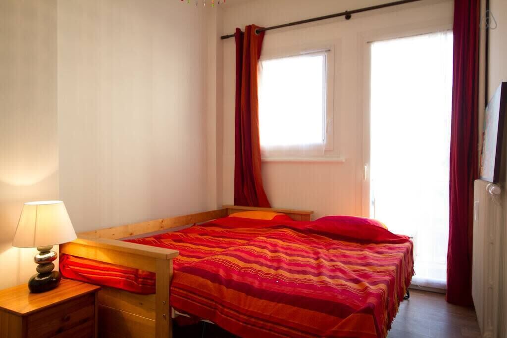 A bedroom with a wooden bed, a nightstand with a lamp, a window with curtains, and a radiator.