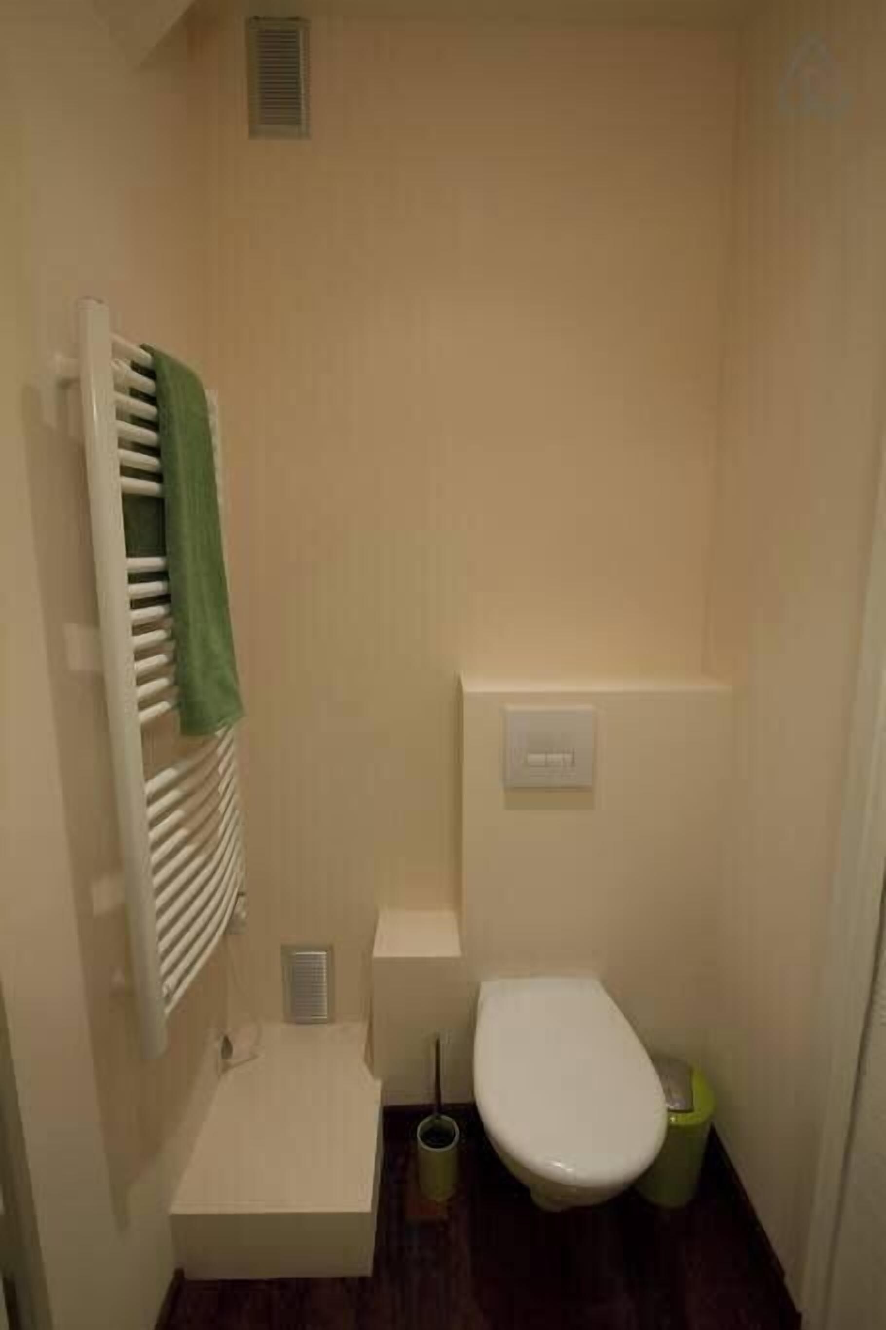 A small, compact bathroom with a white toilet, a green towel hanging on a radiator, and a trash bin.