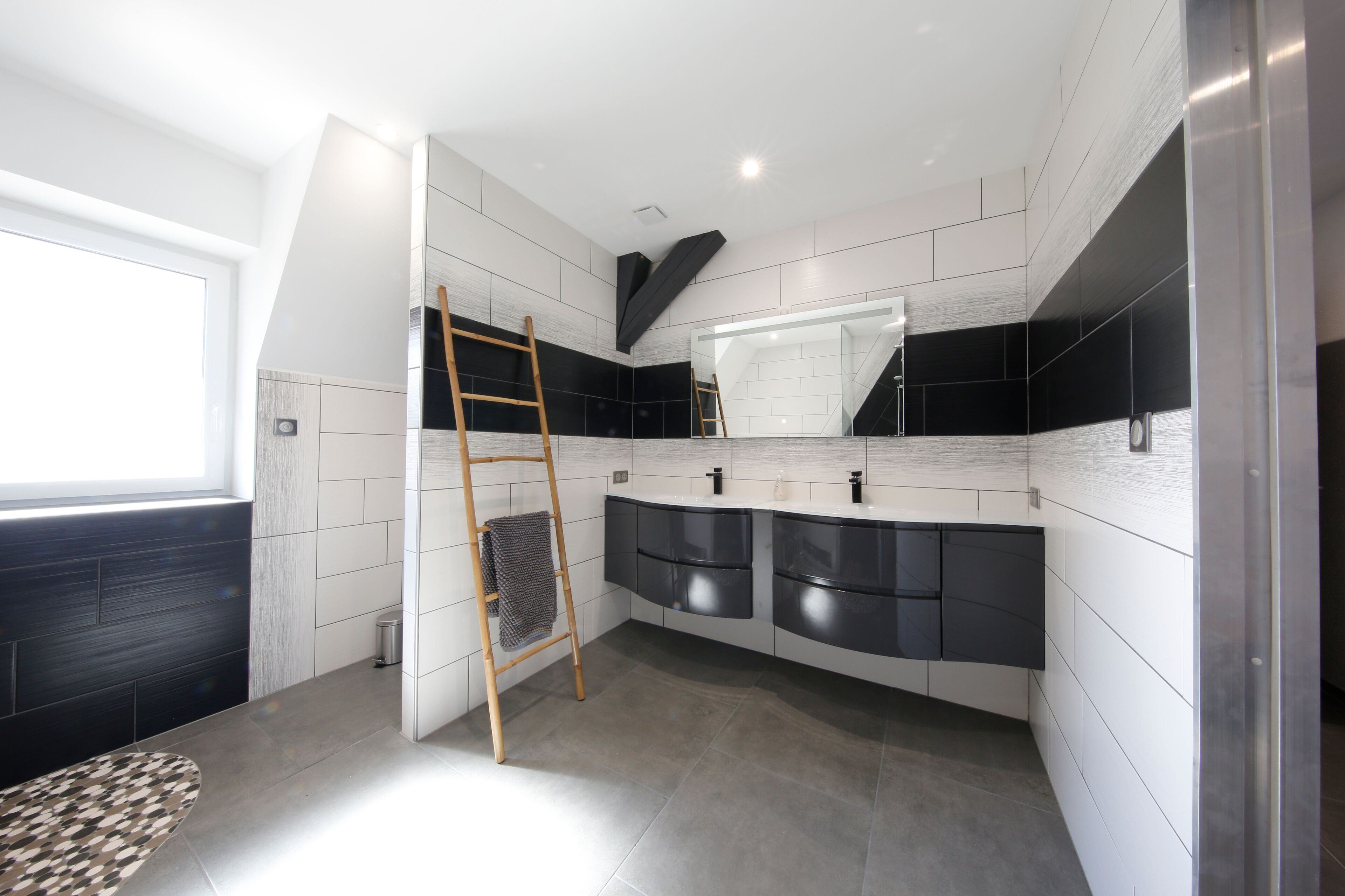 Une salle de bain moderne avec un grand miroir, des murs carrelés aux tons foncés et clairs, une échelle en bois et une fenêtre.