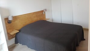 A bedroom with a wooden headboard, a bedside table with a lamp, and a large bed with a dark cover. -  Studio for 2 people in Bages in the heart of the Narbonnaise Regional Natural Park (Bages)