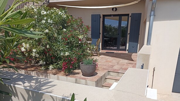 A modern building with a covered entrance, a potted plant, and a small garden area with various plants and flowers. -  Studio for 2 people in Bages in the heart of the Narbonnaise Regional Natural Park (Bages)