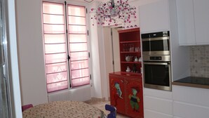 A compact kitchen with a dining area, featuring a refrigerator, oven, and a window with pink blinds. - Superb apartment in the Marais (Paris)