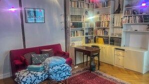 A cozy living room with a red sofa, a patterned armchair, a wooden coffee table, and a red and blue patterned rug. There is a bookshelf filled with books and decorative items, and a small wooden desk with a chair. - Superb apartment in the Marais (Paris)
