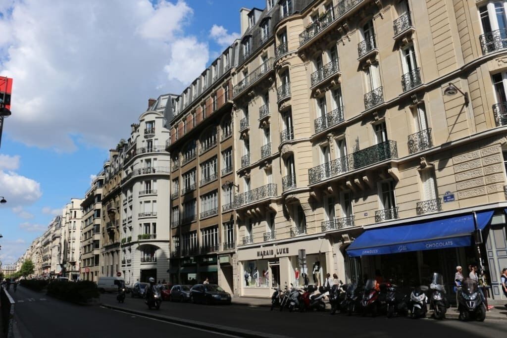 A street with multi-story buildings, a visible storefront with a blue awning, and parked motorcycles.