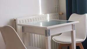 A small room with a white radiator, a white table with a glass top, and two white chairs. There is a blue abstract painting on the wall and a blue curtain on the window. - Meublé de tourisme ** "chezphoenixette (Nieuil)