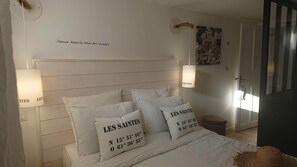 A bedroom with a wooden headboard, white bedding, and pillows with 'LES SAINTES' text. There are two wall-mounted lamps and a door with a striped pattern. - Charming rental T2 Vendée 300m from the beach and 100 m from the forest  (Saint-Jean-de-Monts)