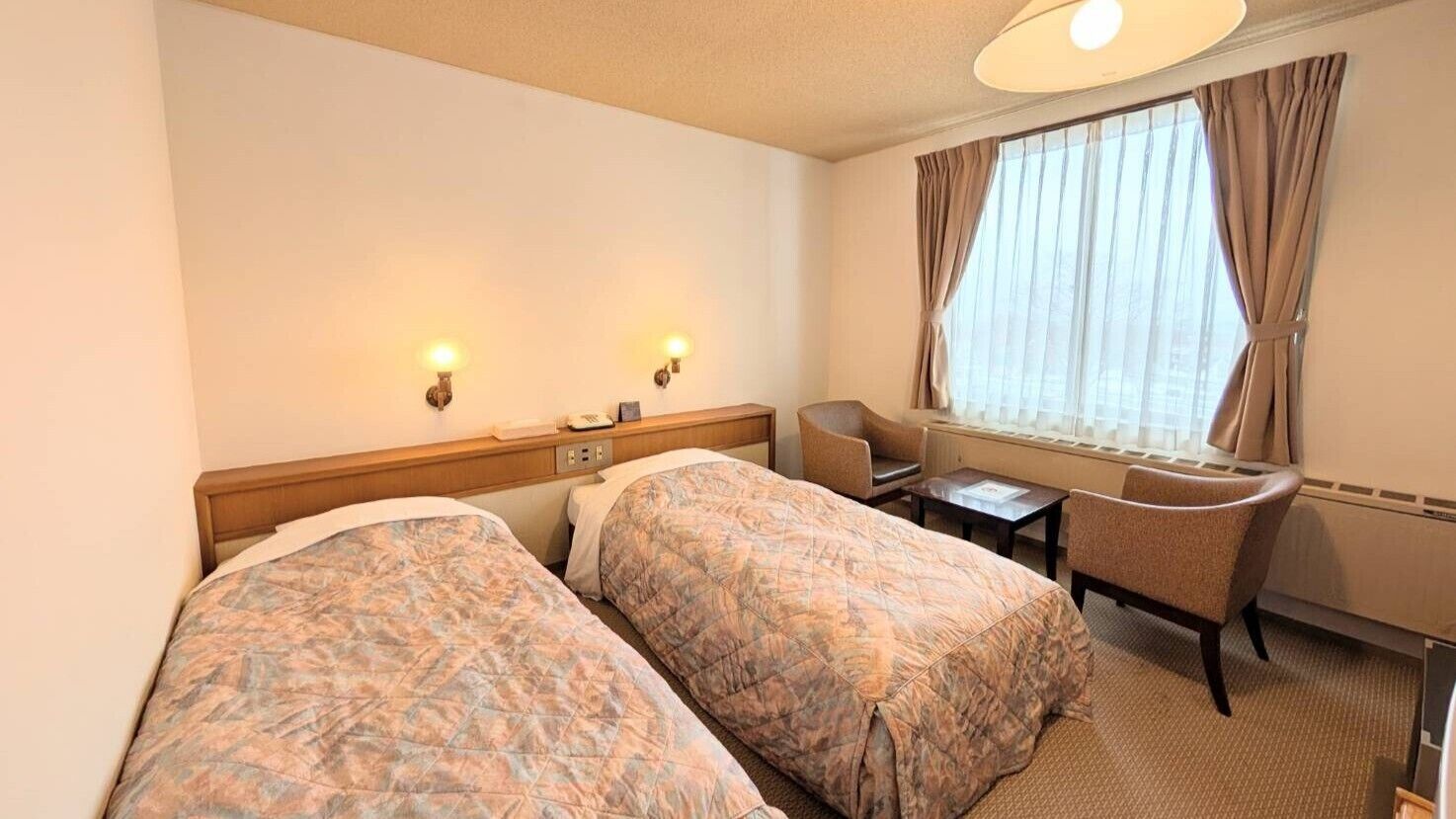 Ein Hotelzimmer mit zwei Betten, einem Schreibtisch, einem Stuhl und einem Fenster mit Vorhängen.
