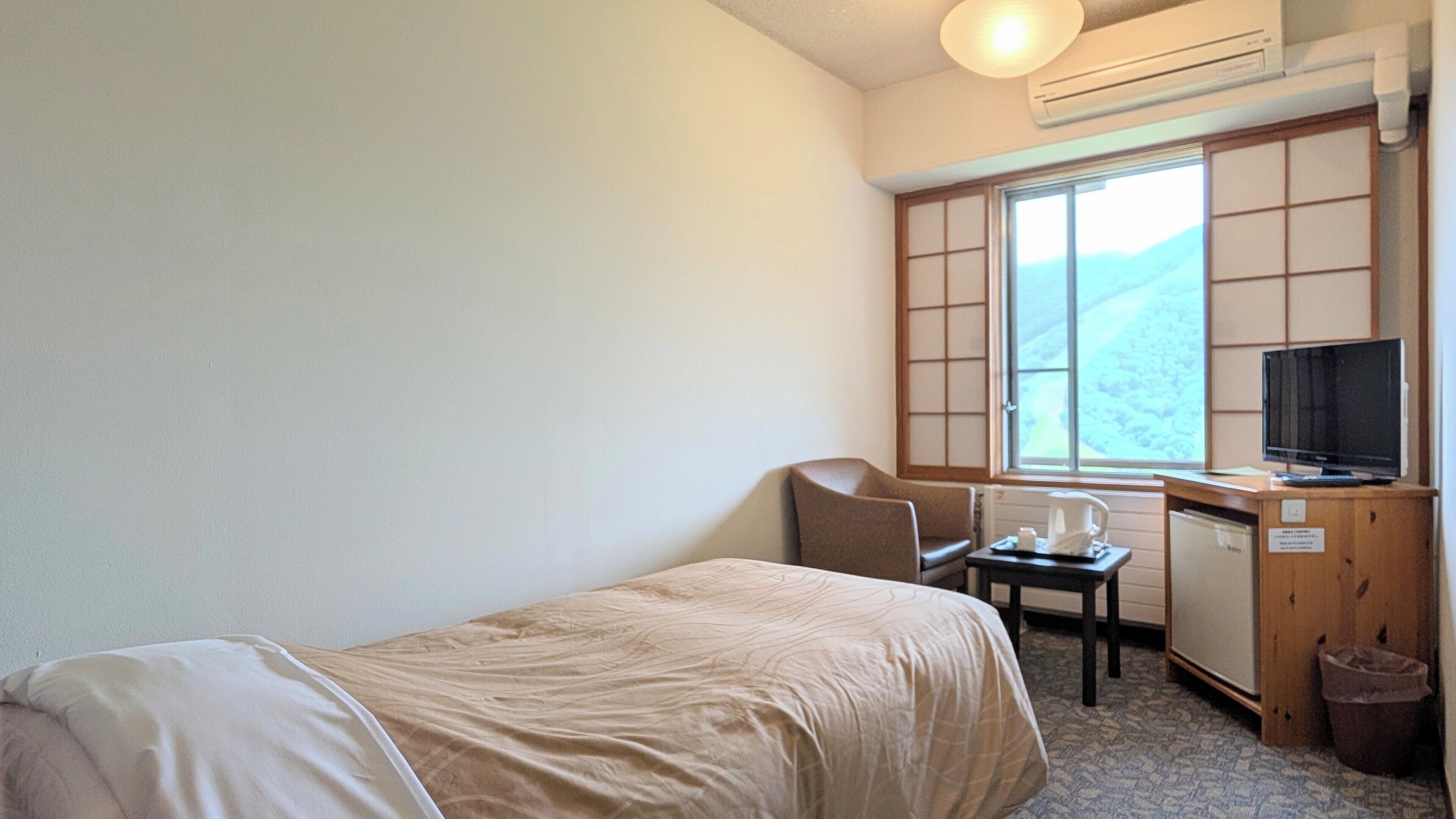 Ein kleines Hotelzimmer mit einem Einzelbett, einem Schreibtisch mit Computer, einem Stuhl, einem Fenster mit Bergblick und einem Fernseher.
