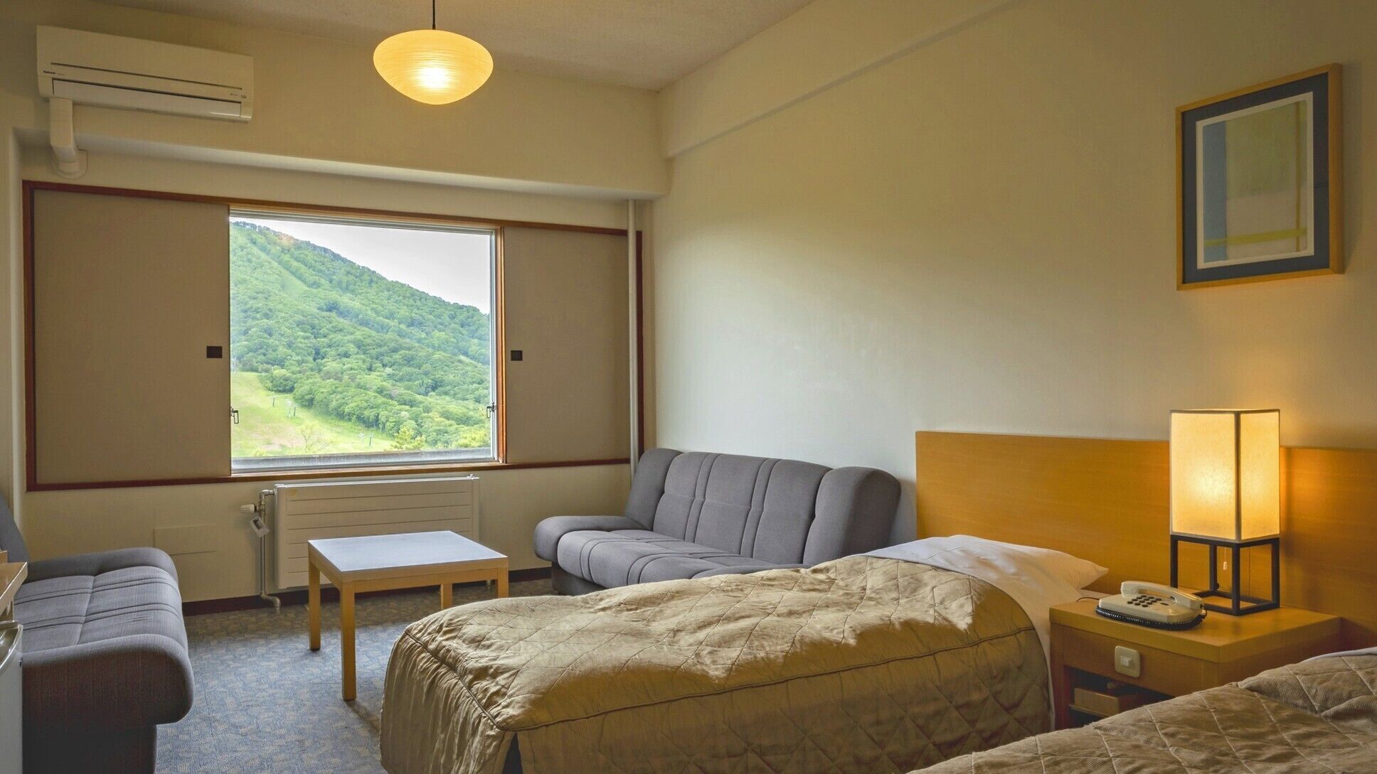 Ein Hotelzimmer mit Bett, Sofa, einem kleinen Tisch, einer Lampe und einem Fenster mit Blick auf eine grüne Landschaft.