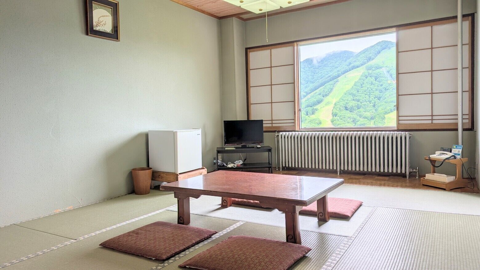 Ein traditioneller japanischer Raum mit Tatami-Boden, einem niedrigen Tisch, einem Fernseher und Blick auf eine gebirgige Landschaft.