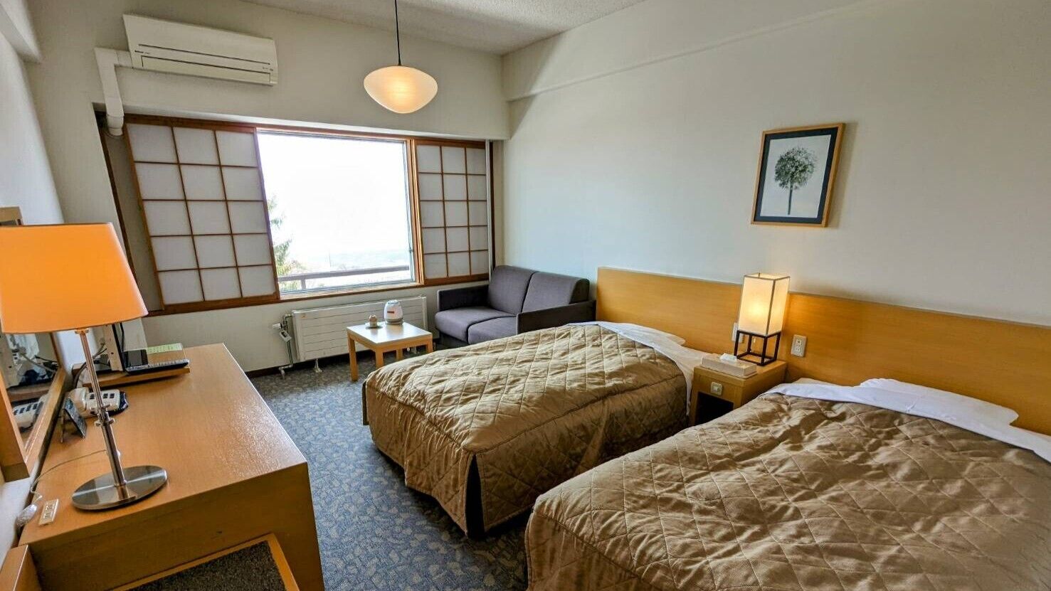 Ein Hotelzimmer mit zwei Betten, einem Schreibtisch mit Lampe, einem Sessel, einem kleinen Tisch, einem Fenster mit Aussicht und einem Wandbild.