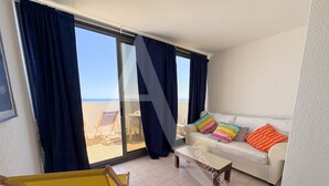 A living room with a sofa, colorful pillows, a small table, a yellow chair, and a large window with curtains.