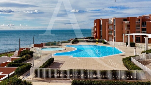 A swimming pool area with multiple pools, surrounded by a building with balconies, and a view of the sea.