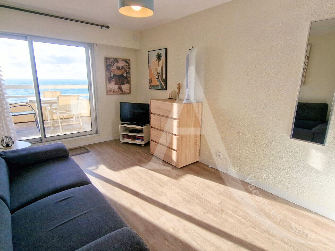 A living room with a sofa, a TV stand, and a wooden cabinet. There is a large window with a view of the ocean.