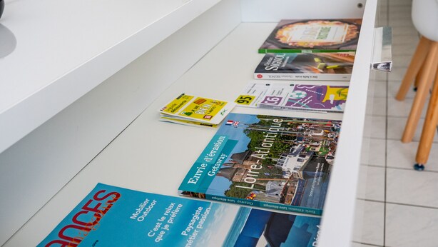 Une cuisine avec un plan de travail blanc, une plante en pot, une lampe et un porte-revues contenant des magazines de voyage.