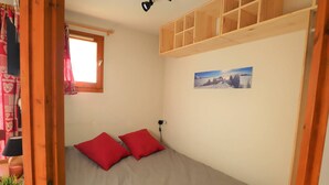 A small room with a bed, two red pillows, a window with curtains, a wall-mounted shelf, and a picture of a snowy landscape.