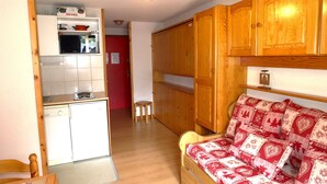 A compact living space with a kitchenette, a red door, wooden cabinets, a patterned sofa, and a small stool. - Cozy apartment for 4 in Arêches-Beaufort, close to ski lifts, shops, and pool (Arêches-Beaufort)