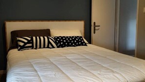 Una cama bien tendida con estructura de madera y ropa de cama blanca, una pared oscura y una puerta a la derecha.