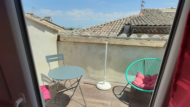 Vista a través de una ventana de un patio con una mesa, sillas, una silla verde y un techo de tejas al fondo.