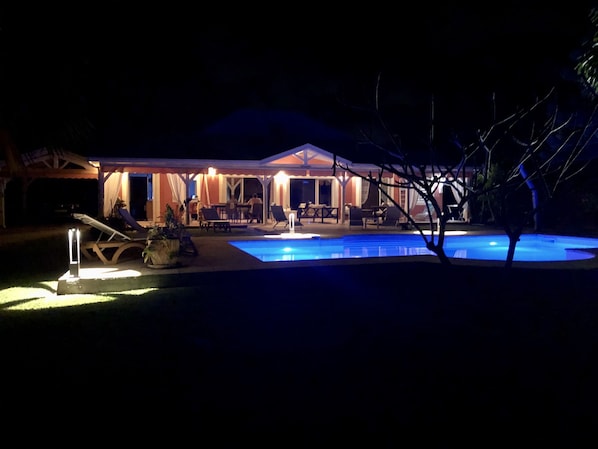 Vista nocturna de una casa con piscina, mobiliario exterior y un patio cubierto.