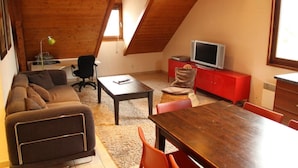 Un espacio tipo loft con techo de madera, un sofá, una mesa de comedor, un televisor sobre un mueble rojo y una ventana que deja entrar luz natural.