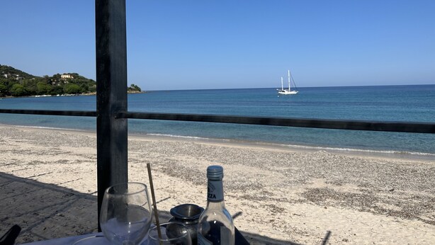 A beachside table with a bottle and glasses, overlooking a calm sea with a sailboat.