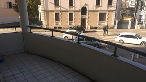 Un balcon doté d’un sol carrelé, d’une rambarde en métal et offrant une vue sur une rue où sont garées des voitures, avec un immeuble en arrière-plan.