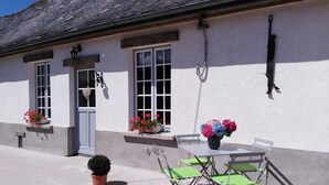 A white building with a grey door, windows with white frames, and a small balcony with green folding chairs and a potted plant. - Welcome to "La Ferme des Tilleuls" (Courset)
