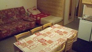 A small, cozy room with a sofa, a single bed, a table with a patterned tablecloth, and a kitchenette with a stove. - RENT APARTMENT IN THE MOUNTAIN - PIERRA MENTA (Villard-Sur-Doron)