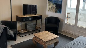 A modern living room with a sofa, a coffee table, a TV on a desk, a painting on the wall, and a view through the window.