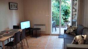 A living room with a TV, a dining table, chairs, a sofa, and a view of a garden through glass doors.