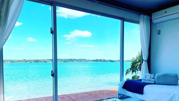 A room with a large sliding glass door leading to a balcony with a view of a calm sea and distant shoreline. - Nel Beach Rosso & Indigo (Nago)