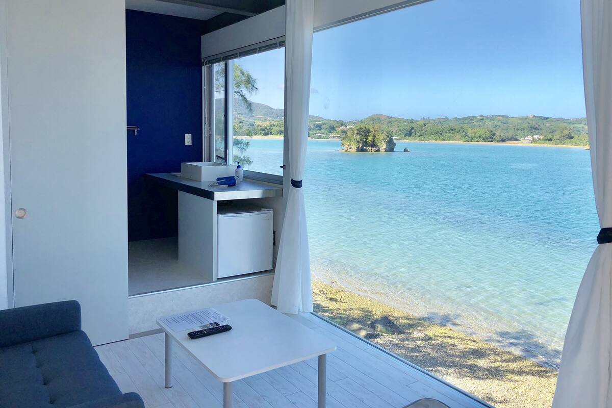 Uma varanda com vista para um lago, uma mesa pequena com um controle remoto e uma área de cozinha compacta.