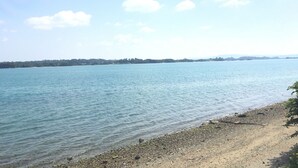 A calm lake with a pebbly shore and distant land. - Nel Beach Rosso & Indigo (Nago)
