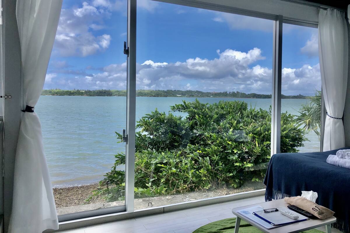 Quarto com uma grande porta de vidro deslizante que dá para uma vista do lago, uma cama com colcha azul escura, uma pequena mesa com um caderno, caneta, controles remotos e uma toalha.