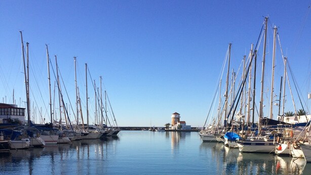 A marina with multiple sailboats docked, calm water, and a small tower structure in the background. - Luxury penthouse overlooking the golf course of Almerimar. (Almerimar)