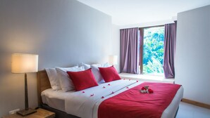 A neatly made bed with red and white bedding, a bedside lamp, and a window with purple curtains. - Residence Bel Eau (Mahé Island)
