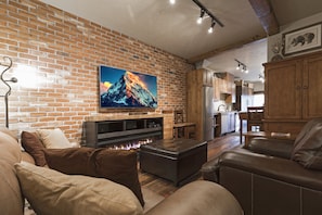 A cozy living room with a brick wall, a fireplace, leather sofas, and a flat-screen TV. - New upgrades to this already brand new studio.  Beautiful Big Mountain views and right downtown! (Whitefish)
