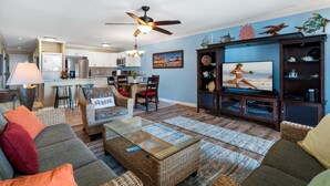Una sala de estar con televisor, muebles de mimbre y una cocina al fondo.