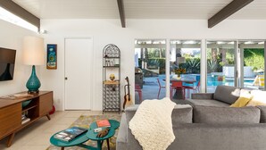 Ein modernes Wohnzimmer mit grauem Sofa, einem türkisfarbenen Couchtisch und einem Bücherregal. Ein großes Fenster bietet Blick auf eine Außenpatio mit Pool und Gartenmöbeln.