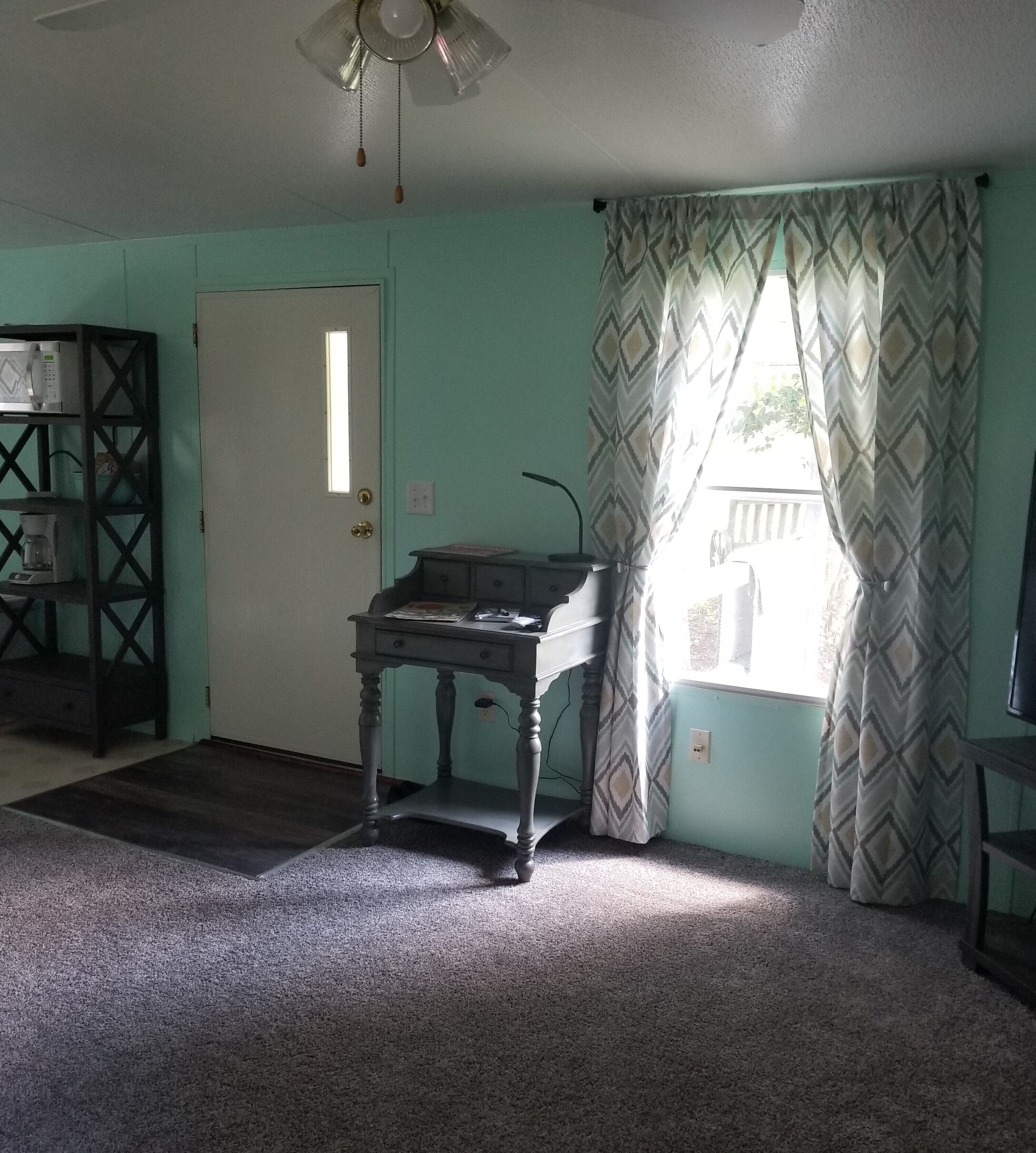 Una habitación con escritorio, ventilador de techo, puerta y ventana con cortinas estampadas.