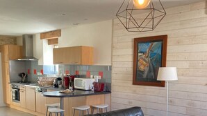 A modern living room with a black leather sofa, a dining area with stools, a kitchen with wooden cabinets, and a painting on the wall. - Gîte de Penn Ar Roz: 3 épis seaside GUILER-SUR-GOYEN, 4-5 pers, swimming pool. (Guiler-sur-Goyen)