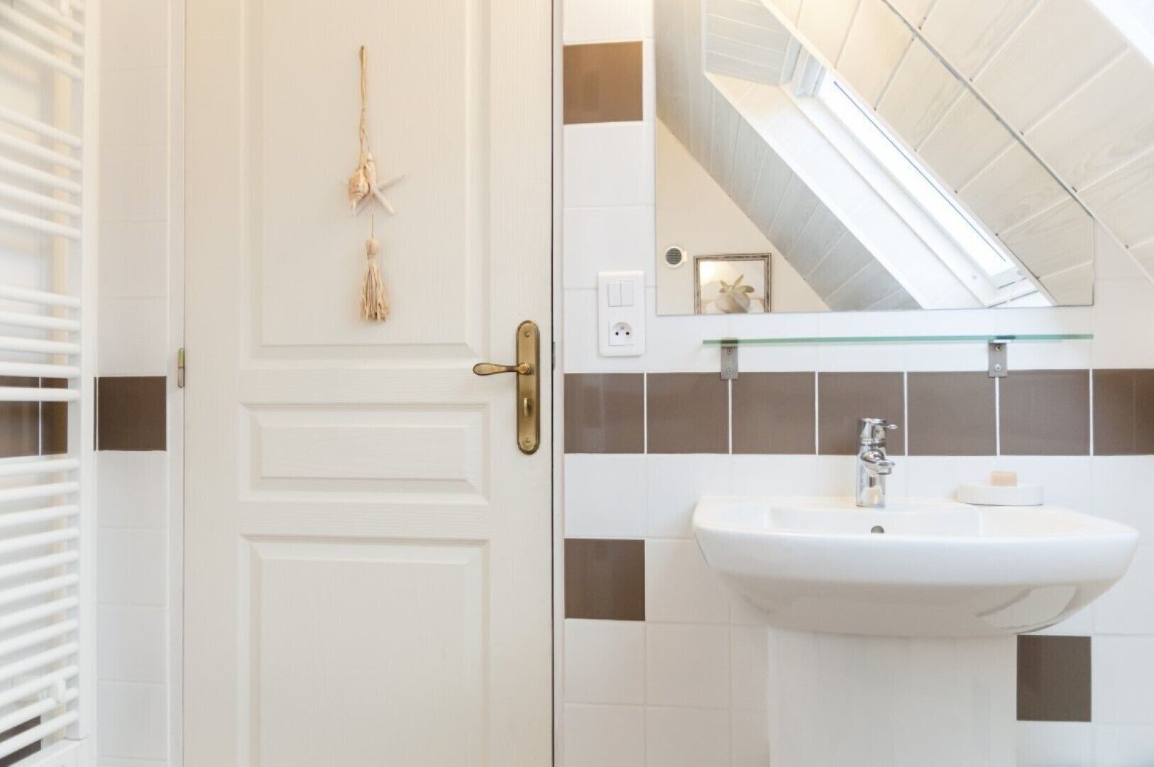 Une salle de bain avec une porte blanche, une étagère en verre au-dessus d’un lavabo blanc et un miroir qui reflète un escalier.